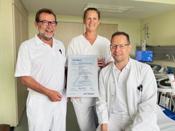 Zwei Ärzte und eine Schwester halten das Zertifikat und lächeln.